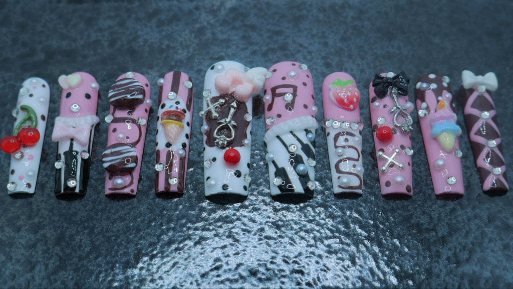 Ten decorative false nails arranged in a row on a dark, textured surface. The nails feature pink, white, and black candy-themed designs with small red cherry accents and rhinestone embellishments.